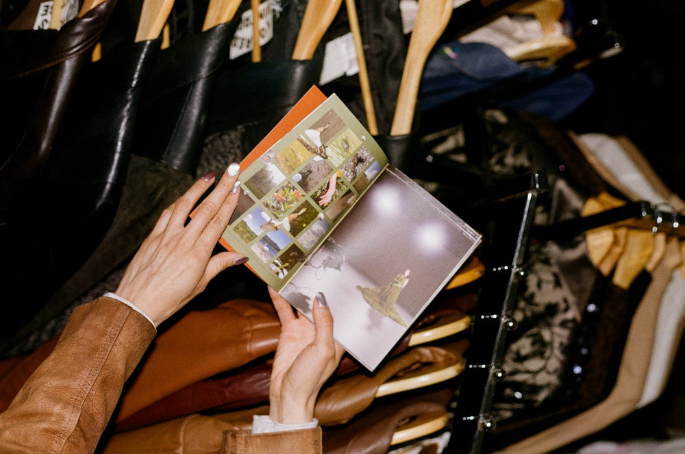 A person in a brown jacket holds open a Congrats Records fashion lookbook featuring recent projects with models. Behind them, garments hang on wooden hangers, suggesting ties to the creative community.