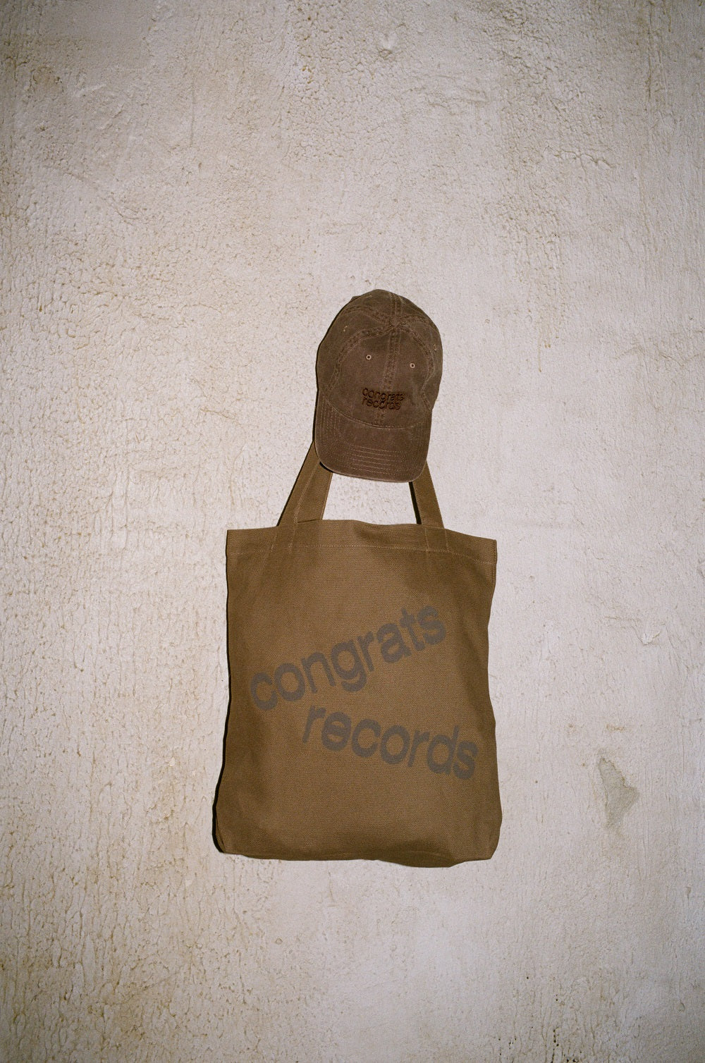 A brown Congrats Records tote bag with the brand name in gray and a matching brown cap hang on a textured off-white wall, highlighting the stylish appeal of Congrats Records accessories.