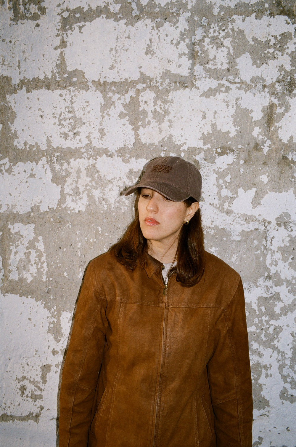A person in a brown leather jacket and gray cap, featuring Congrats Records, stands before a rough white brick wall, looking down with a neutral expression.
