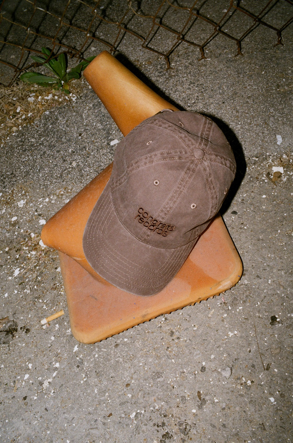 A Congrats Records brown baseball cap sits atop two orange traffic cones on cracked pavement near a chain-link fence, surrounded by small weeds and scattered debris.