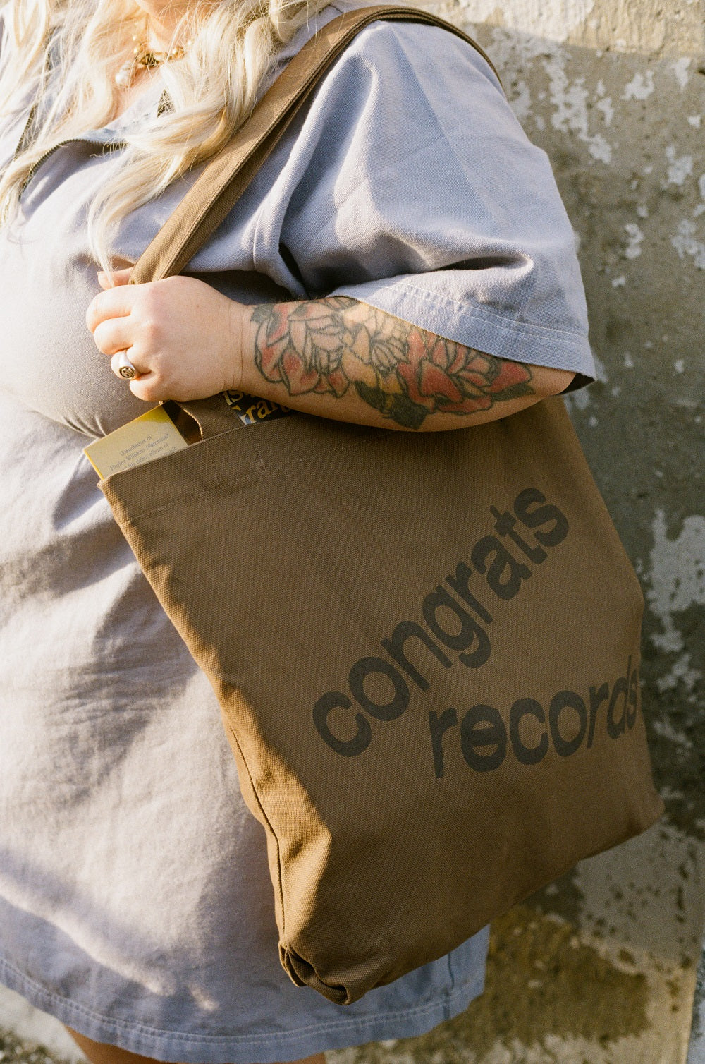 A person with long, wavy, light hair and a tattooed arm holds a brown Congrats Records Logo Tote Bag with black text, paired with a loose-fitting, light gray shirt.