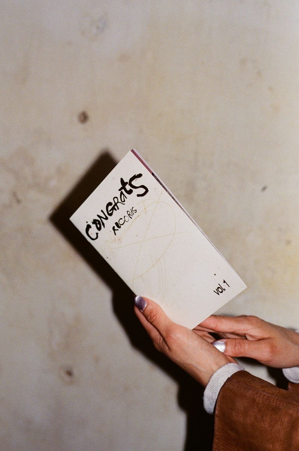 A person with painted nails holds a Congrats Records zine titled CONCERTS RECORDS vol. 1 against a beige textured wall, reflecting their connection with the creative community.