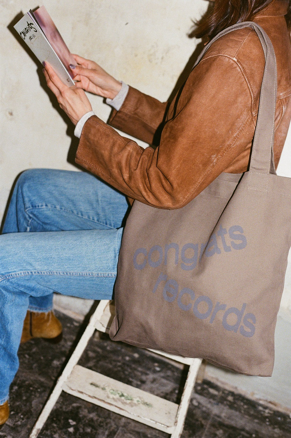 Wearing a brown jacket and blue jeans, a person reads on a white stool with a large brown Congrats Records tote bag—signaling their creative spirit and possible involvement with the brand’s projects or zines.